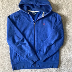 Blå hoodie från Polo Ralph Lauren - Säljer en blå hoodie från Polo Ralph Lauren med dragkedja och huva. Tröjan har en röd broderad logga på bröstet och är tillverkad i mjuk bomull. Perfekt för en avslappnad stil och enkel att matcha med jeans eller joggers.