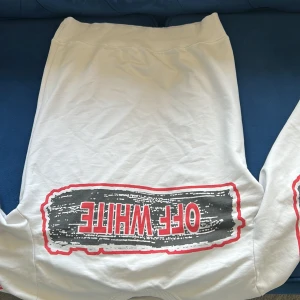 Vit hoodie från Off-White - Säljer en hoddie från Off-White med stora svarta och röda tryck på både bak- och framsidan. Hoodien har elastisk midja och Off-White-logga samt pilar på baksidan. Perfekta för en streetwear-look.