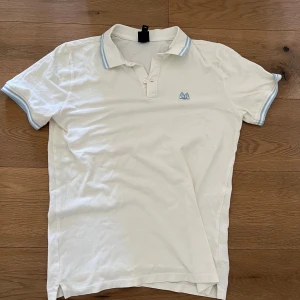 Vit pikétröja med blå detaljer från HM - Vit pikétröja med ljusblå ränder på krage och ärmslut. Liten broderad logga på bröstet. Klassisk pikékrage och knappar framtill. Perfekt för en avslappnad stil.