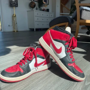 Nike Air Jordan 1 Mid röd/svart/vit - Säljer ett par Nike Air Jordan 1 Mid i klassisk färgkombination av rött, svart och vitt. Skorna har högt skaft, röd swoosh och snörning framtill. Perfekta för dig som gillar streetwear och sneakers med ikonisk design.