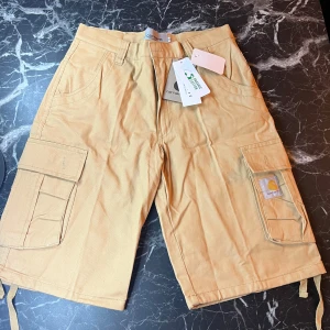 Beige cargoshorts från Carhartt - Snygga beige cargoshorts från Carhartt med flera praktiska fickor på sidorna. Klassisk loose fit och robust bomullsmaterial, perfekta för sommardagar. Stora fickor med lock och Carhartt-logga på benet ger en cool och funktionell look.
