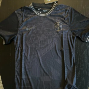 Svart Brasilien fotbollströja Nike - Snygg svart Brasilien fotbollströja från Nike med diskret mönster och broderat klubbmärke på bröstet. Tröjan har korta ärmar, rund hals med randig ribbad kant och är tillverkad i lätt, ventilerande funktionsmaterial.