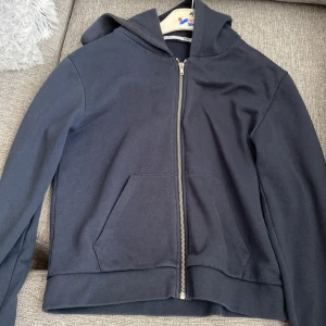 Mörkblå hoodie med dragkedja, S - Mörkblå hoodie i storlek S med hel dragkedja framtill och huva. Tröjan har inte andvända många gånger! Tröjan har mjukt fleecefoder på insidan och en stor ficka framtill. Perfekt för kyliga dagar och enkel att matcha med jeans eller joggers.