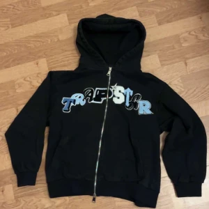 Trapstar kofta  - Svart hoodie från Trapstar med stor logga i blått, vitt och svart på bröstet. Tröjan har huva, dragkedja framtill och två fickor. Perfekt för dig som gillar streetwear och vill sticka ut med en fet design.diskutera pris ✨✨