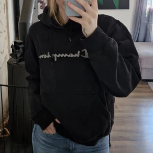 Svart hoodie från Champion - Säljer en svart hoodie från Champion med broderad logga över bröstet. Tröjan har huva med dragsko och en stor magficka. Perfekt för dig som gillar en avslappnad och klassisk stil. Fläck på ena ärmen (baksidan) enligt bild.