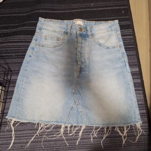Ljusblå jeanskjol med fransad kant - Säljer en ljusblå jeanskjol från Perfect Jeans med fransad nederkant och klassiska fickor både fram och bak. Kjolen har en rak passform och är i kort modell, perfekt till sommaren. Stängs med dragkedja och knapp framtill.