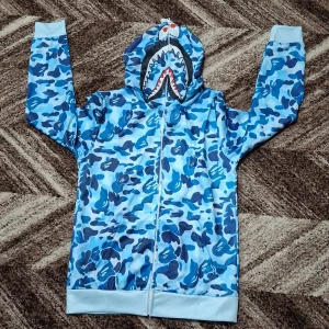 Blå camo hoodie med hajmotiv - Säljer en blå camouflagemönstrad hoodie med dragkedja och hajmotiv på huvan. Tröjan har långa ärmar, vita muddar och detaljer i orange och gul på huvan. Perfekt för dig som gillar streetwear och unika plagg.