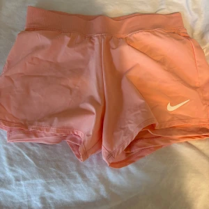 Nike ljusrosa träningsshorts - Snygga ljusrosa träningsshorts från Nike med elastisk midja och klassisk vit swoosh-logga på ena benet. Lätta och luftiga i syntetmaterial, perfekta för gymmet eller löprundan. Sportig look och skön passform.
