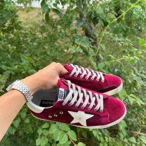 Vinröda Golden Goose sneakers mocka - Snygga vinröda sneakers från Golden Goose i mjuk mocka med vit stjärndetalj på sidan och vita snören. Klassisk låg modell med platt sula och svartvit logga på plösen. Perfekta för dig som vill sticka ut med stilrena detaljer.