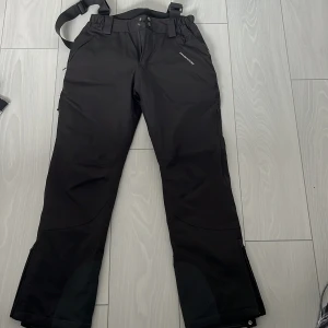 Svarta täckbyxor Swedemount strl 36 - Svarta täckbyxor från Swedemount med justerbara hängslen och förstärkta benslut. Byxorna har dragkedjor vid bensluten och är gjorda i slitstarkt syntetmaterial, perfekta för vinterns alla äventyr. Snygg och funktionell design med praktiska detaljer.
