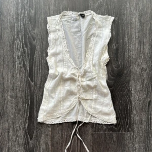 Vit spetsväst med snörning från InMorocco - Supersöt vit väst i bomull från InMorocco med broderade detaljer och spetskant längs kanterna. Västen har v-ringning och knytning framtill för en romantisk vibe. Snygg till Jeans 