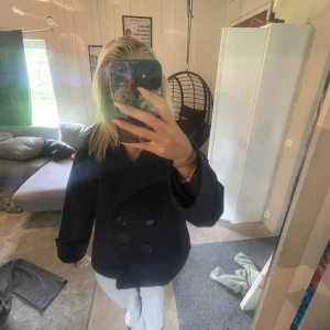 Kappa design by si - Säljer en svart kappa med dubbelknäppning och bred krage. Jackan har en klassisk, rak passform och är tillverkad i ett tjockare ullmaterial som håller dig varm under kyliga dagar. Perfekt för dig som gillar en stilren och tidlös look.