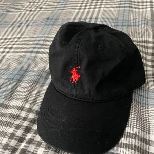 Svart keps från Polo Ralph Lauren - Svart keps från Polo Ralph Lauren med klassisk röd broderad logga framtill och 'POLO' broderat bak. Justerbar rem baktill med metallspänne. Snygg och tidlös modell i bomull som passar till allt.