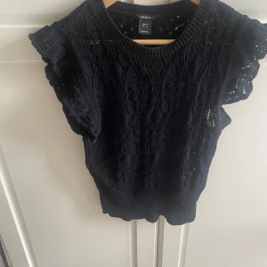 Svart stickad topp från Lindex S - Supersöt svart stickad topp från Lindex i storlek S. Toppen har ett genombrutet mönster, volangliknande ärmar och ribbade kanter. Perfekt för dig som gillar en feminin och trendig look med snygga detaljer.