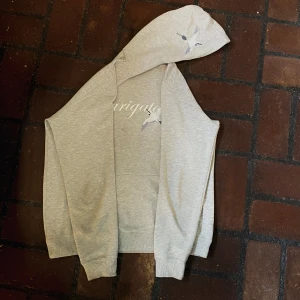 Hoodie från Axel Arigato  - Snygg ljusgrå hoodie från Axel Arigato med broderad fågel och texten 'arigato' på bröstet. Tröjan har huva och långa ärmar, tillverkad i mjuk bomullsmix. Perfekt för en chill och stilren look.