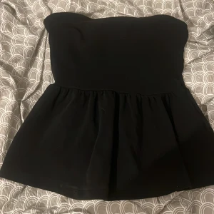 Svart tubtopp från Gina Tricot - Supersnygg svart tubtopp från Gina Tricot i peplum-modell. Toppen är axelbandslös och har en utsvängd nederdel som ger en cool och trendig siluett. Perfekt att styla med jeans eller kjol för en chic look.