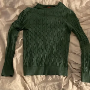 Grön kabelstickad tröja Morris XS - Snygg grön kabelstickad tröja från Morris i 100% merinoull. Tröjan har lång ärm, rund halsringning och en broderad logga på bröstet. Perfekt för dig som gillar klassisk stil med en modern touch.