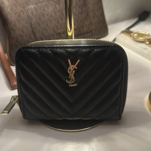 Svart quiltad plånbok från Saint Laurent - Snygg svart plånbok från Saint Laurent i quiltat skinn med guldiga detaljer och dragkedja runtom. Klassisk YSL-logga framtill och flera fack för kort och sedlar. Perfekt storlek för att ha med sig överallt och ger en lyxig känsla.