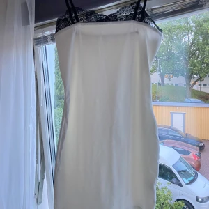 Vit klänning med spetsdetalj - Säljer en vit kort klänning med tunna axelband och svart spets upptill. Klänningen har en slimmad passform och en stilren, minimalistisk look.