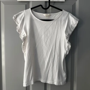 Vit volangtopp med spets från H&M - Söt vit topp från H&M med volangdetaljer och spets längs ärmarna. Rund halsringning och ärmlös design som ger en luftig känsla. 