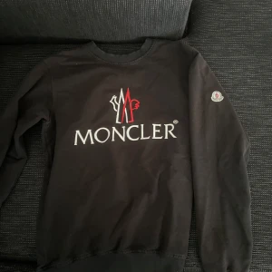 Svart sweatshirt från Moncler - Svart sweatshirt från Moncler med stort broderat logotyptryck i vitt och rött på bröstet samt Moncler-märke på ena ärmen. Tröjan har rund halsringning, långa ärmar och ribbade muddar vid ärmslut och nederkant. Perfekt för dig som gillar stilrena märkesplagg.