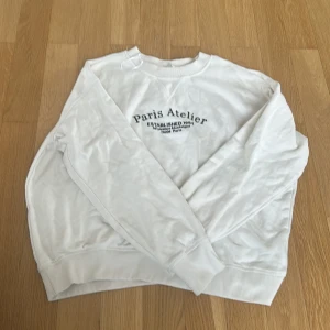 Vit sweatshirt Paris Atelier S - Vit sweatshirt från Paris Atelier med svart texttryck på bröstet. Tröjan har rund halsringning, långa ärmar och ribbade muddar vid ärmslut och nederkant. Perfekt för en clean och avslappnad look.