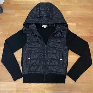 Svart cardigan från EE Eligance Essentials - Svart cardigan med quiltad framsida och huva. Ärmar och rygg är stickade för en skön kontrast och extra komfort. Jackan har dragkedja framtill och två fickor med tryckknappar. Perfekt för kyliga dagar när du vill ha både stil och värme.