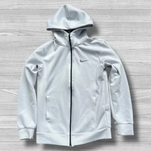 Vit Nike vindjacka med huva - Snygg vit vindjacka från Nike med svart dragkedja och klassisk swoosh-logga på bröstet. Jackan har huva, två fickor och ribbade muddar. Perfekt för dig som gillar sportig stil och vill ha något lätt att slänga på sig.