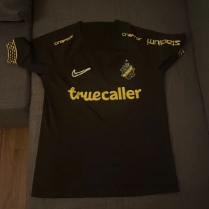 AIK svart matchtröja Nike - Snygg svart AIK fotbollströja från Nike med gula detaljer och tryck. Truecaller-logga på bröstet, klubbmärke och sponsortryck på ärmarna. Kortärmad modell i lätt och ventilerande material, perfekt för match eller träning.
