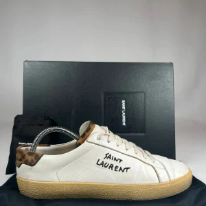 Saint Laurent vita sneakers med leoparddetalj - Saint Laurent sneakers i vitt skinn med handskriven logga på sidan och cool leopardmönstrad detalj vid hälen. Klassisk rund tå, beige gummisula och snörning framtill. Perfekta för dig som vill ha en unik touch på dina sneakers.