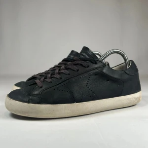 Svarta sneakers från Golden Goose - Svarta sneakers från Golden Goose med klassisk låg profil, snörning och vit platt sula. Skorna är i skinn med diskret stjärndetalj i perforerat mönster på sidan och texten GCDB/STAR i guld. Perfekta för en clean och stilren look. Utan box