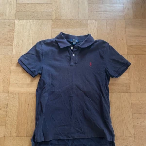 Mörkblå pikétröja från Ralph Lauren - Klassisk mörkblå pikétröja från Polo Ralph Lauren med röd broderad logga på bröstet. Tröjan har korta ärmar, krage och två knappar framtill. Perfekt för en clean och stilren look. Tillverkad i mjuk bomull för skön känsla hela dagen.