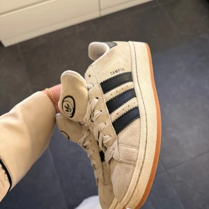 Beige adidas Campus sneakers mocka - Snygga adidas Campus sneakers i beige mocka med klassiska svarta ränder på sidorna och gummisula i brun ton. Skorna har vit snörning och rund tå, samt adidas-logga på plösen. Perfekta för dig som gillar retrostil och streetwear.