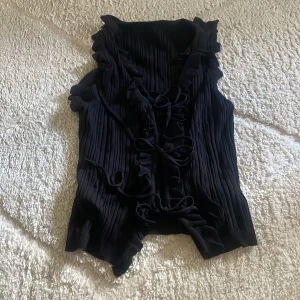 Svart plisserad kofta med volang - Svart väst med plisserat tyg och volangdetaljer längs kanterna. Västen har snörning framtill och är ärmlös. 