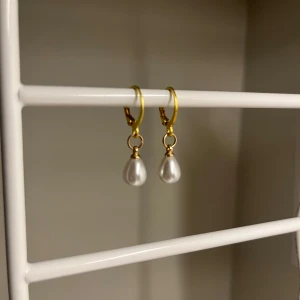 Guldiga örhängen med pärldroppar - Stainless Steel. Snygga örhängen med guldfärgade hoops och droppformade vita pärlor. Perfekta för att ge en elegant touch till din look. Klassisk design som passar till många stilar.