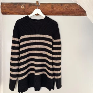 Randig stickad tröja från Isolde XL - Snygg stickad tröja från Isolde i svart med breda beige ränder. Oversized känsla med lång ärm och rund halsringning. Perfekt för dig som gillar en chill och klassisk look med lite edge. Passar till jeans eller kjol för en enkel stil.