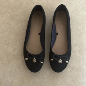 Svarta ballerinaskor Tommy Hilfiger - Stilrena svarta ballerinaskor från Tommy Hilfiger i mocka-look med rund tå. Dekorativa rosetter framtill med guldfärgade detaljer och TH-logga. Snyggt mönstrat innerfoder och platt sula för en klassisk vibe.