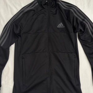 Svart Adidas vindjacka med dragkedja och byxor - Svart vindjacka från Adidas med klassiska grå ränder längs ärmarna och diskret logga på bröstet. Jackan har hel dragkedja framtill, ståkrage och två fickor. Tillverkad i polyester, perfekt för sportiga vibes eller chill dagar. MED BYXOR!