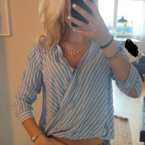 Blåvit omlottblus med ränder - Snygg blå skjorta med vita ränder och omlottdesign. Skjortan har lång ärm och v-ringad halsringning, perfekt för en trendig och avslappnad look. Passformen är normal och materialet känns lätt och bekvämt.