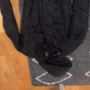 Svart quiltad jacka Vinson Polo Club - Svart quiltad jacka från Vinson Polo Club med huva och dragkedja. Jackan har snygga detaljer som broderad logga på bröstet och två dragkedjefickor framtill. Materialet är lätt och smidigt, perfekt för kyligare dagar.