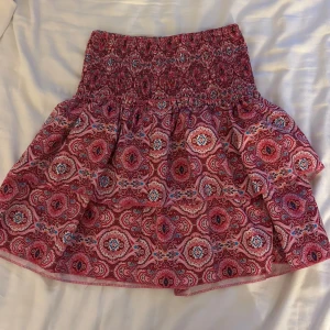 Rosa mönstrad volangkjol - Supersöt kort kjol i rosa med färgglatt mönster och bred smock i midjan. Kjolen har volanger som ger extra fladder och en riktigt somrig vibe. (Säljer en likadan i blå)