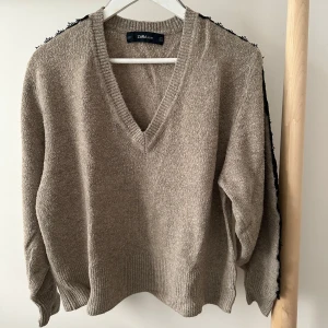 Beige stickad tröja med spets från Zara - Beige stickad tröja från Zara med v-ringning och långa ärmar. Snygg detalj med svart spets längs axlar och ärmar som ger en unik touch. Mjuk och skön. Ribbstickade muddar vid ärmslut och nederkant.
