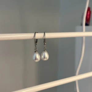 Silvriga örhängen med vita pärlor -  I rostfritt stål, handgjord. Snygga örhängen med droppformade vita pärlor och silvriga detaljer. Perfekta för att ge en elegant touch till din look. Enkla att bära tack vare smidiga krokar. Klassisk design som aldrig går ur tiden.
