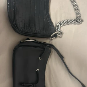 1 för 30 båda för 50 - Säljer två svarta axelväskor. Ena har croco-mönster och en chunky silverkedja som axelrem, medan den andra har slät yta och cool snörningsdetalj framtill. Båda är i syntetmaterial och har trendig, stilren look som passar till många outfits.
