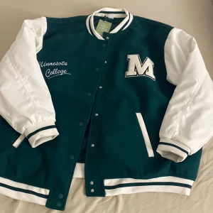 Grön och vit college bomberjacka - Storlek är M. Snygg bomberjacka i grönt och vitt med broderad text 'Minnesota College' och stor M-patch framtill. Jackan har vita ärmar, grönt tyg på kroppen, randiga muddar och knappar framtill. Perfekt för en sportig och trendig look. 