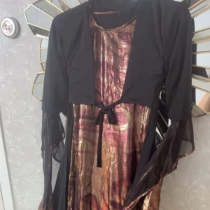 Svart rödrosa kurdisk klänning  - Unik svart kurdisk klänning med glittriga, skimrande inslag i guld och rödrosa toner framtill. Klänningen har långa, vida ärmar med transparent tyg. Perfekt för dig som vill sticka ut!✨Passar barn från 6-8 år✨