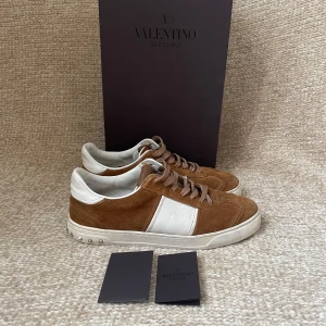 Valentino Garavani bruna sneakers mocka - Snygga sneakers från Valentino Garavani i brunt mocka med vita skinnpaneler och vita detaljer. Klassisk låg modell med snörning och platt sula. Perfekta för dig som vill ha en stilren och lyxig look med en twist.