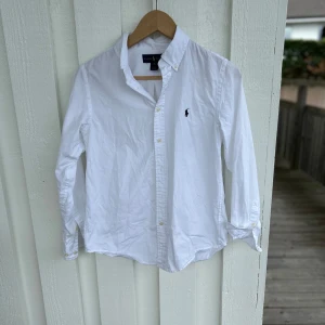 Vit skjorta från Ralph Lauren - Klassisk vit skjorta från Ralph Lauren med broderad logga på bröstet. Skjortan har lång ärm, knappar framtill och en snygg krage. Tillverkad i bomull och har en clean, tidlös look som funkar till allt. Custom fit-modell för en snygg passform.