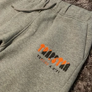 Gråa Tripstar mjukisbyxor med brodyr - Snygga grå mjukisbyxor från Tripstar med broderad logga i orange och svart på benet. Byxorna har dragsko i midjan, fickor och smal passform. Perfekta för chill eller streetwear. Materialet är mjukt och skönt, troligtvis bomull eller polyester.