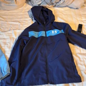 Blå vindjacka från Under Armour - Snygg marinblå vindjacka från Under Armour med ljusblå detalj över bröstet och vit logga. Jackan har huva, dragkedja framtill och två sidofickor. Perfekt för dig som gillar sportig stil och vill ha något lätt och smidigt till vardagen.